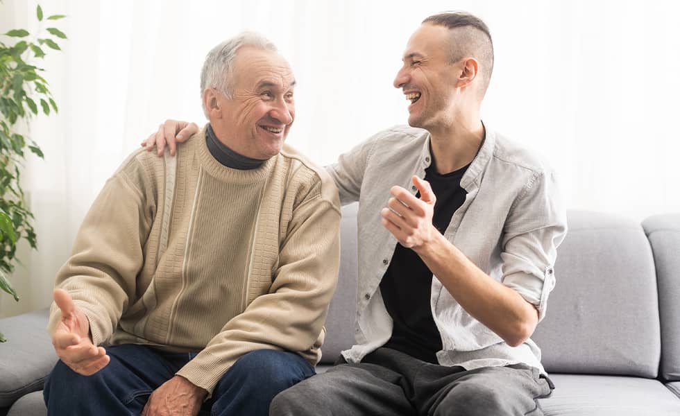 Adult son laughing with his father while sitting on the couch together
