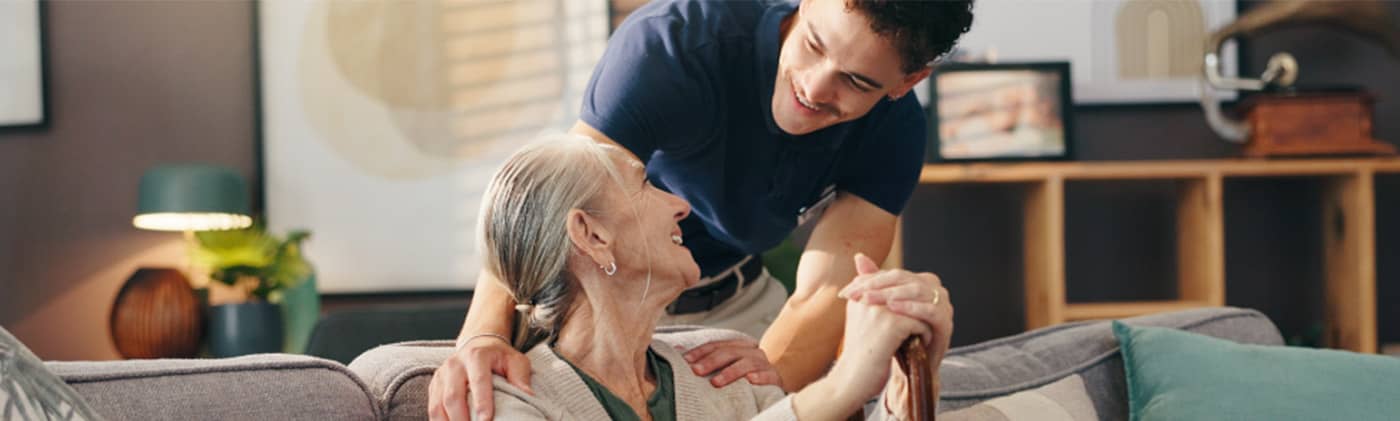 A young caregiver in a navy shirt smiles warmly at an elderly woman seated on a couch in a cozy living room.