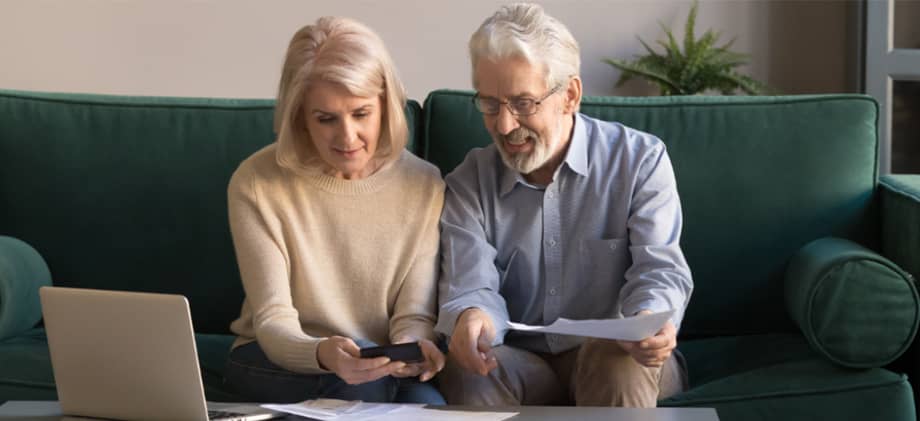 Elderly couple sitting on a couch reviewing financial documents with a laptop and calculator.