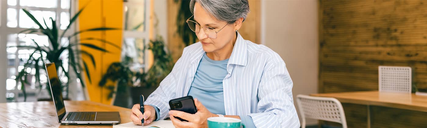 Older woman sitting at a table, writing in a notebook while looking at her phone, with a laptop beside her.