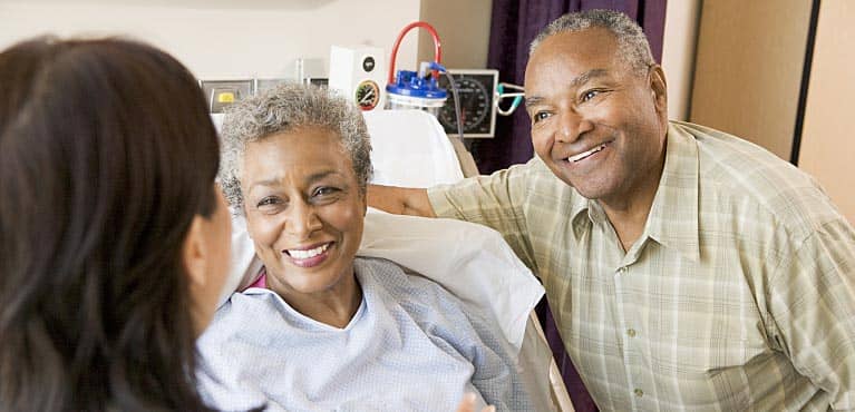 husband embraces wife in hospital bed as they consult a doctor and discuss living benefits together