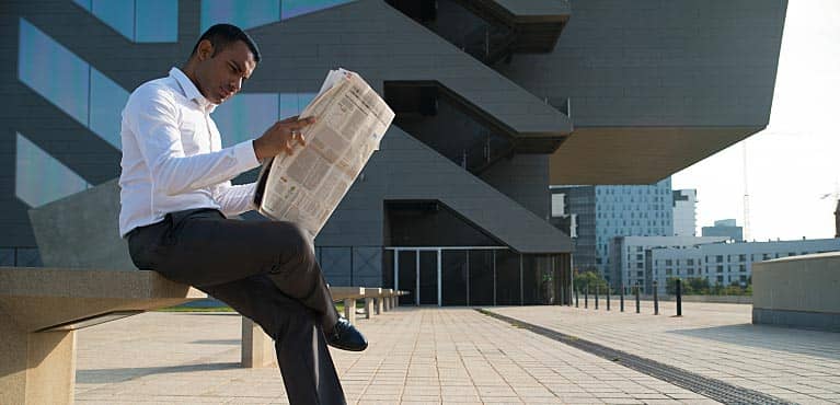 businessman sits outside office and checks performance of mutual funds vs. stocks in newspaper