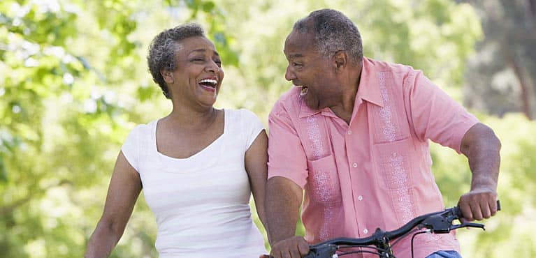 older couple laughs as they ride bikes down a path and discuss whole life insurance together
